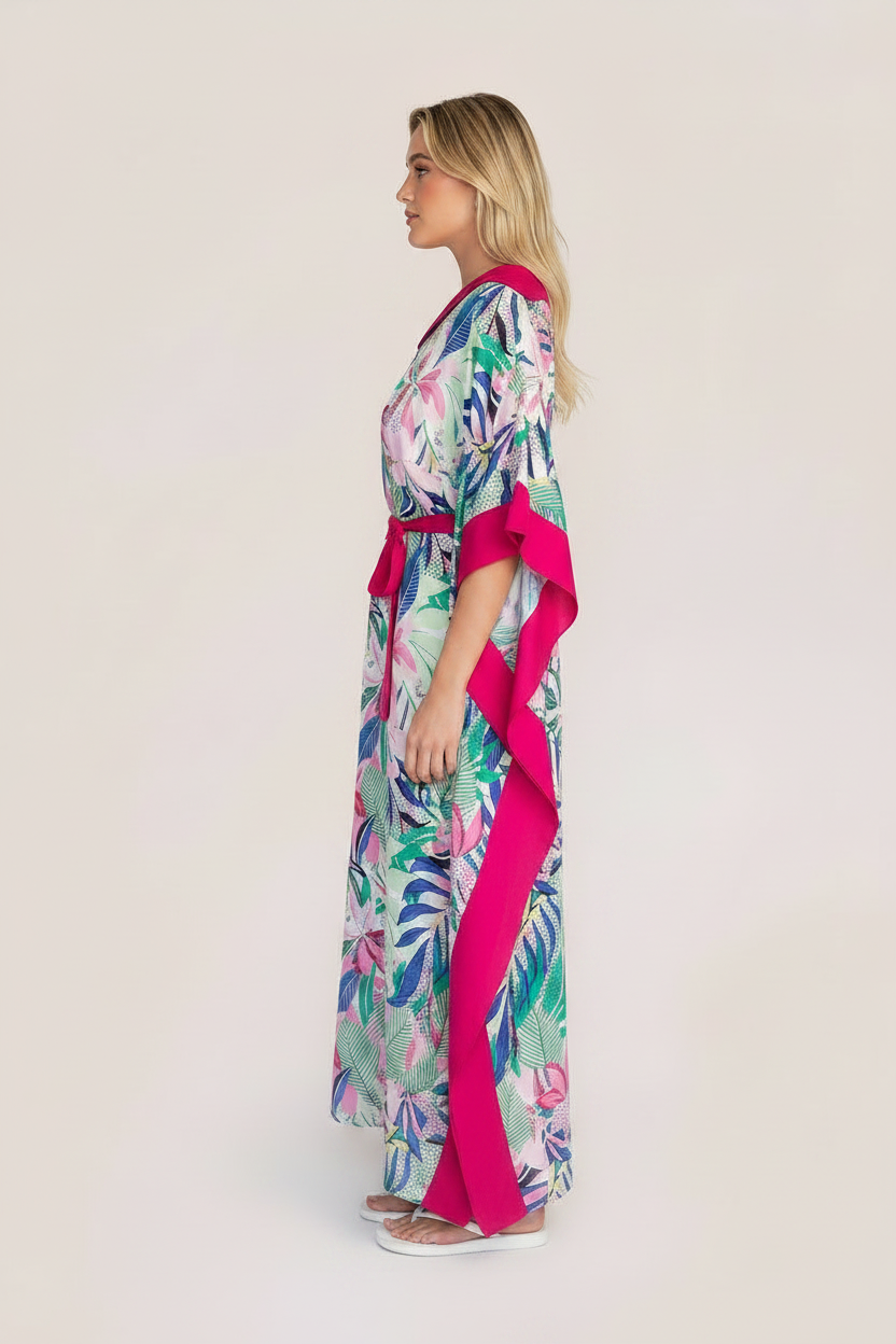 A woman modeling a tropical print kaftan robe with a vibrant pink border and sash, set against a plain light beige studio background.