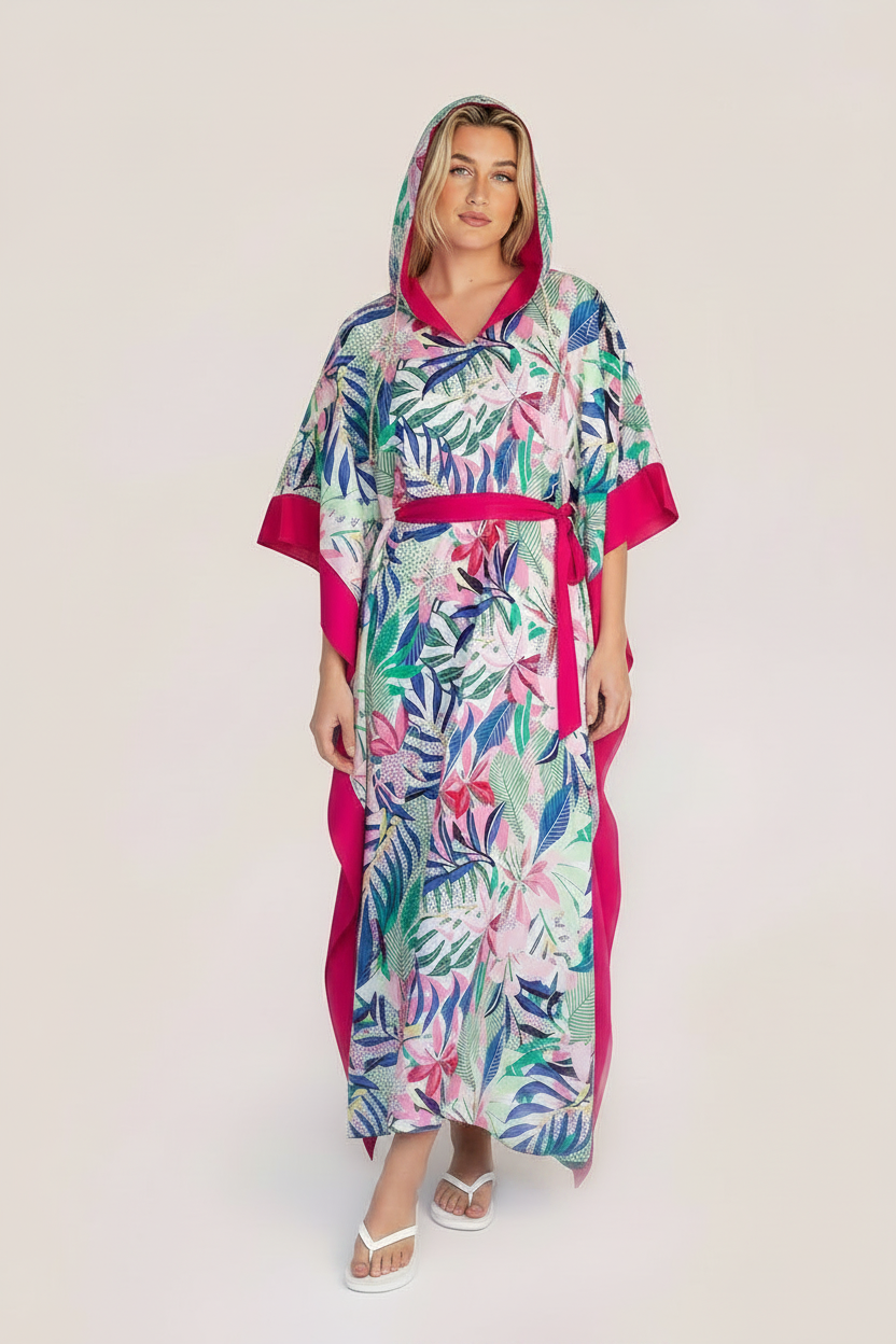 A woman modeling a tropical print kaftan robe with a vibrant pink border and sash, set against a plain light beige studio background.
