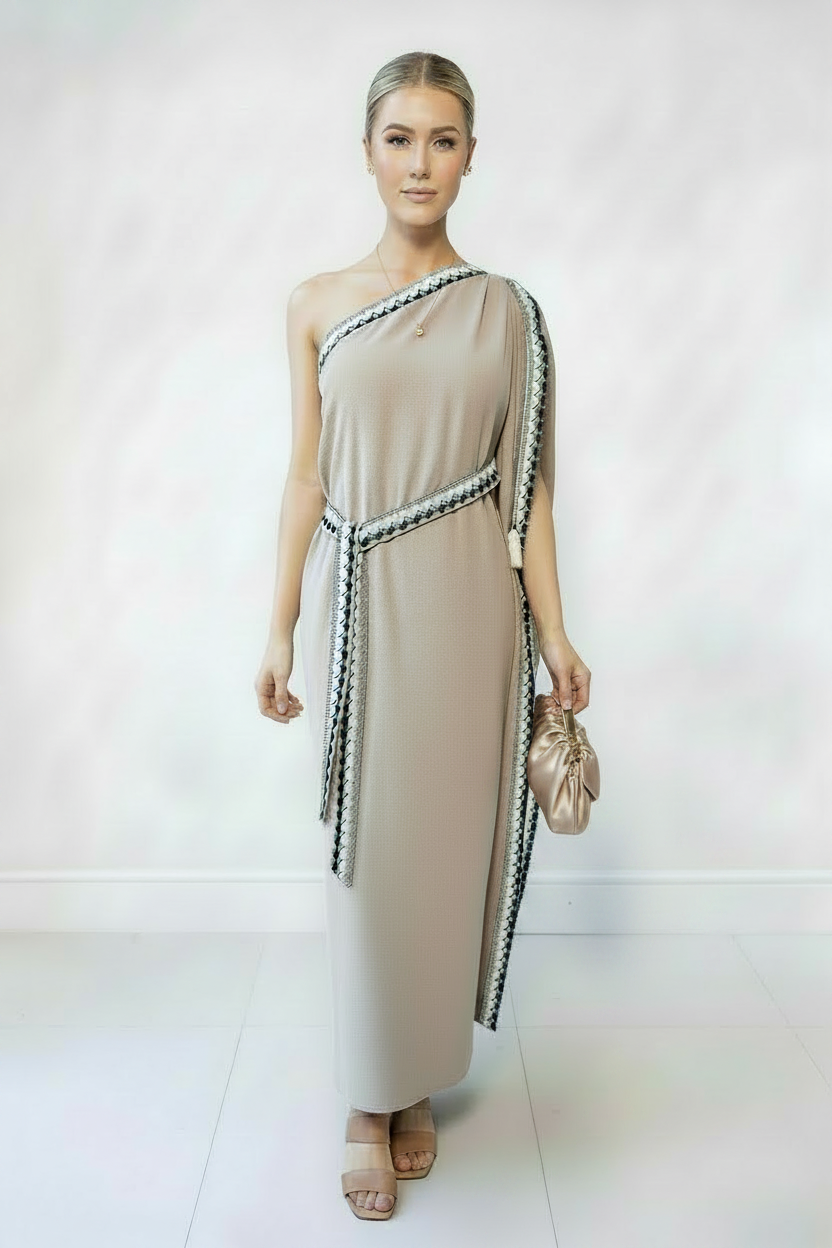 A woman modeling a beige one-shoulder maxi dress with a black and beige patterned draped scarf, holding a metallic gold clutch bag against a light gray studio background.
