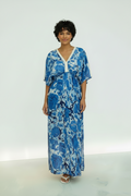 A woman modeling a blue and white floral kaftan dress with a V-neckline and short sleeves, standing against a clean white studio background.