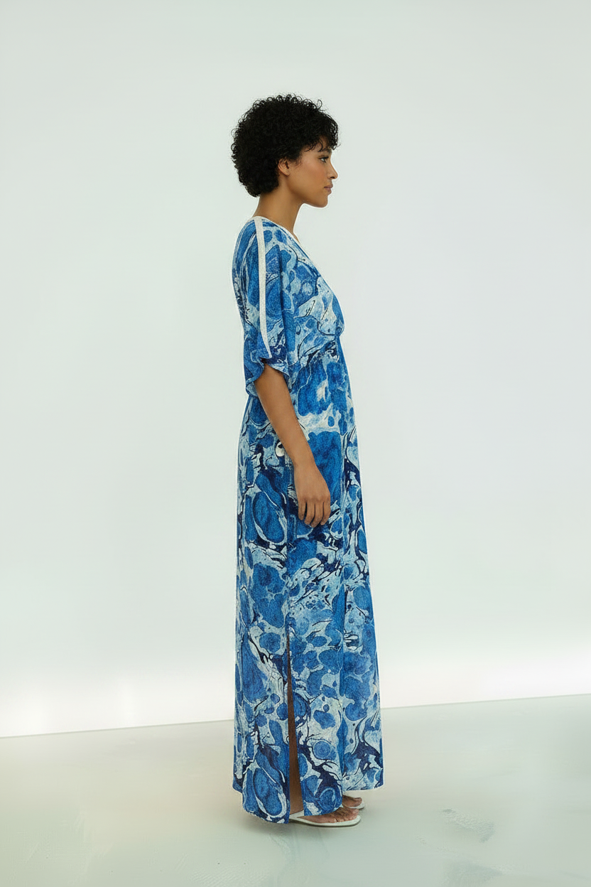 A woman stands in profile wearing a long blue and white abstract print maxi dress with elbow-length sleeves and a side slit, photographed against a clean white studio background.