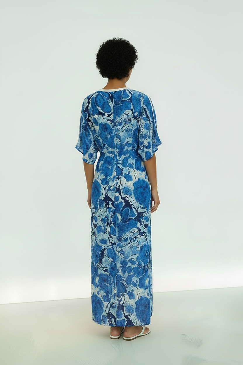 A woman stands in profile wearing a long blue and white abstract print maxi dress with elbow-length sleeves and a side slit, photographed against a clean white studio background.