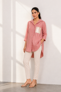 A woman models a dusty pink linen shirt with a textured metallic pocket, paired with off-white trousers and beige sandals, standing against a white wall with soft window shadows.