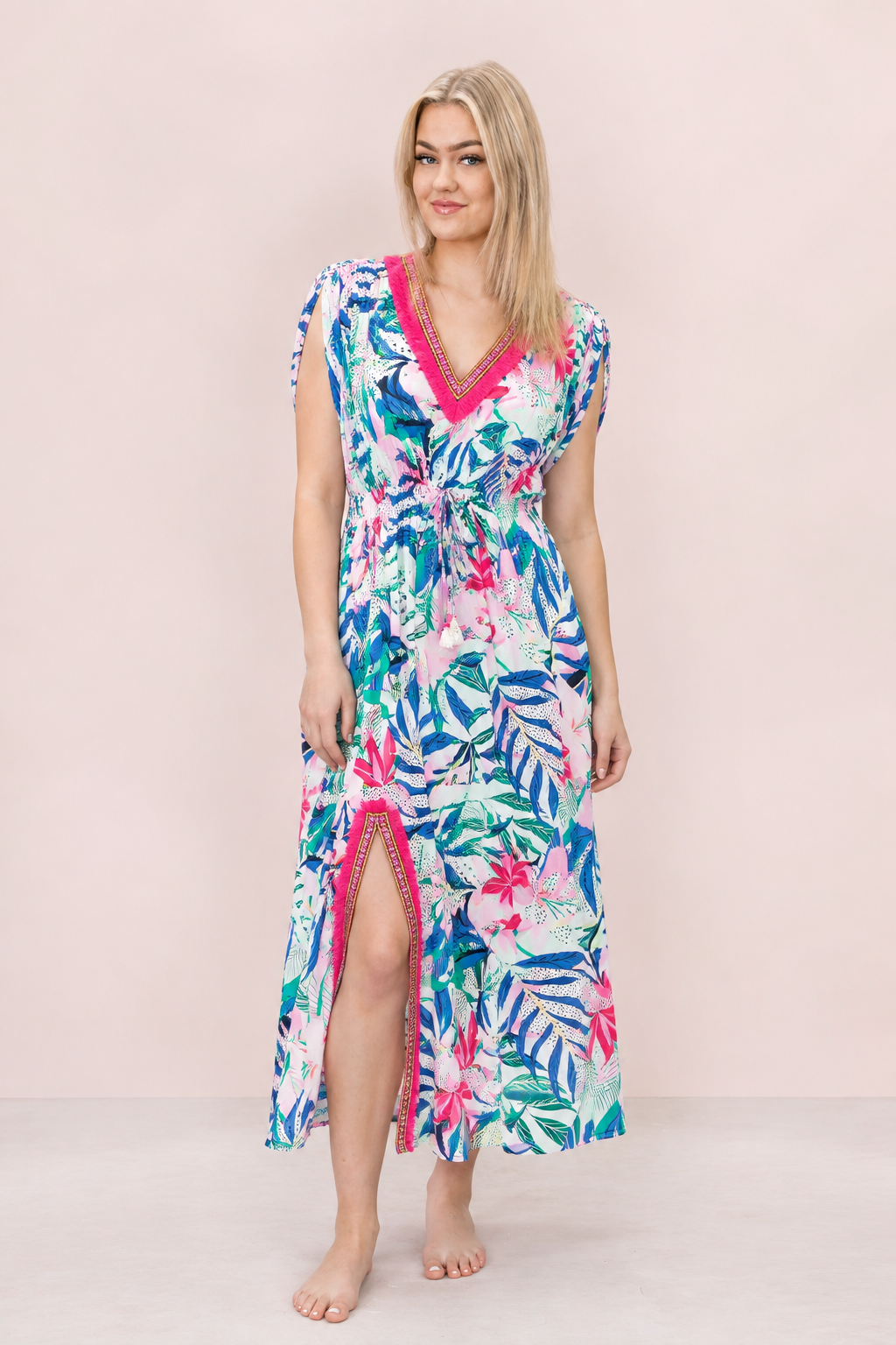 A woman modeling a tropical print midi dress with a pink trim, standing against a solid pale pink background.