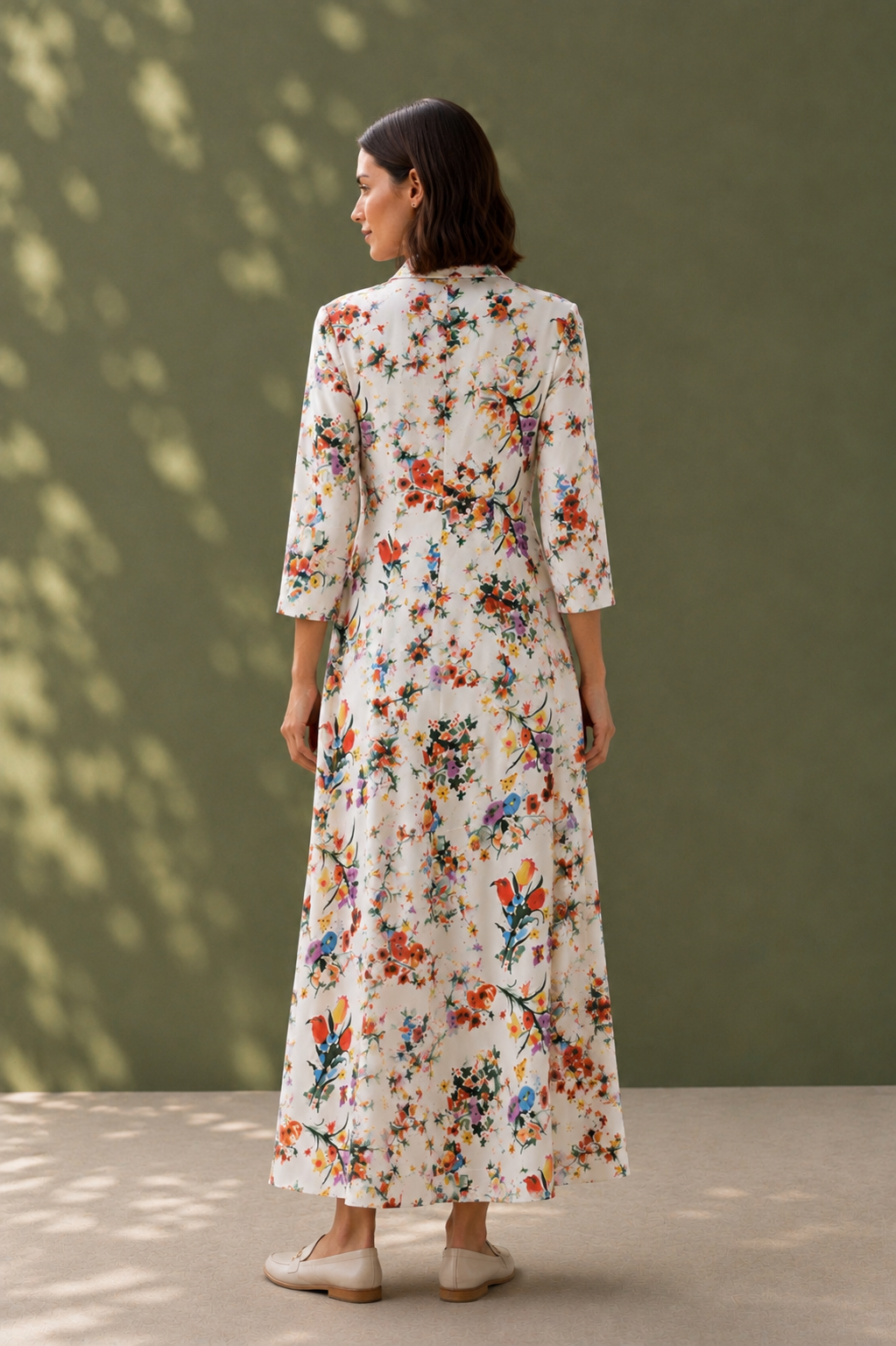 A woman stands with her back to the camera wearing a long-sleeved, cream-colored maxi dress with a colorful floral and bird pattern, set against a muted olive green wall with soft leaf shadows.
