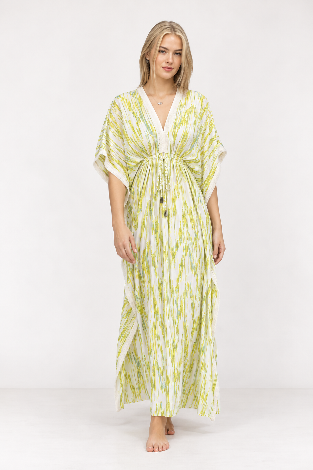 A woman modeling a long kaftan-style dress with a white base and lime green vertical brushstroke pattern, featuring a V-neckline, drawstring waist, and short wide sleeves, set against a plain white studio background.