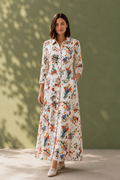 A woman stands outdoors against a muted green wall with soft leaf shadows, wearing a long-sleeve white maxi shirt dress covered in a colorful floral and bird print. The dress has a button front, collar, and side pockets, paired with light beige shoes.