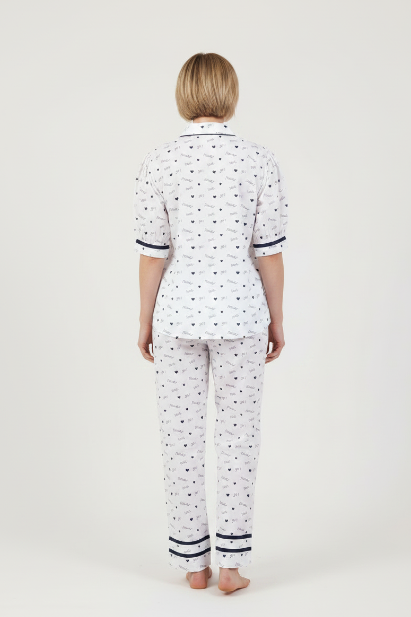 A woman modeling a two-piece pajama set featuring a white base with a small navy blue pattern, a short-sleeve button-up top, and matching long pants, set against a plain light gray studio background.
