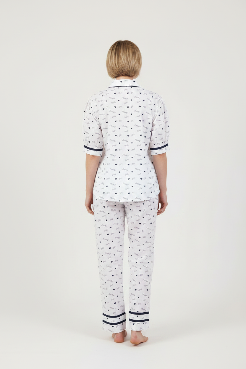 A woman modeling a two-piece pajama set featuring a white base with a small navy blue pattern, a short-sleeve button-up top, and matching long pants, set against a plain light gray studio background.