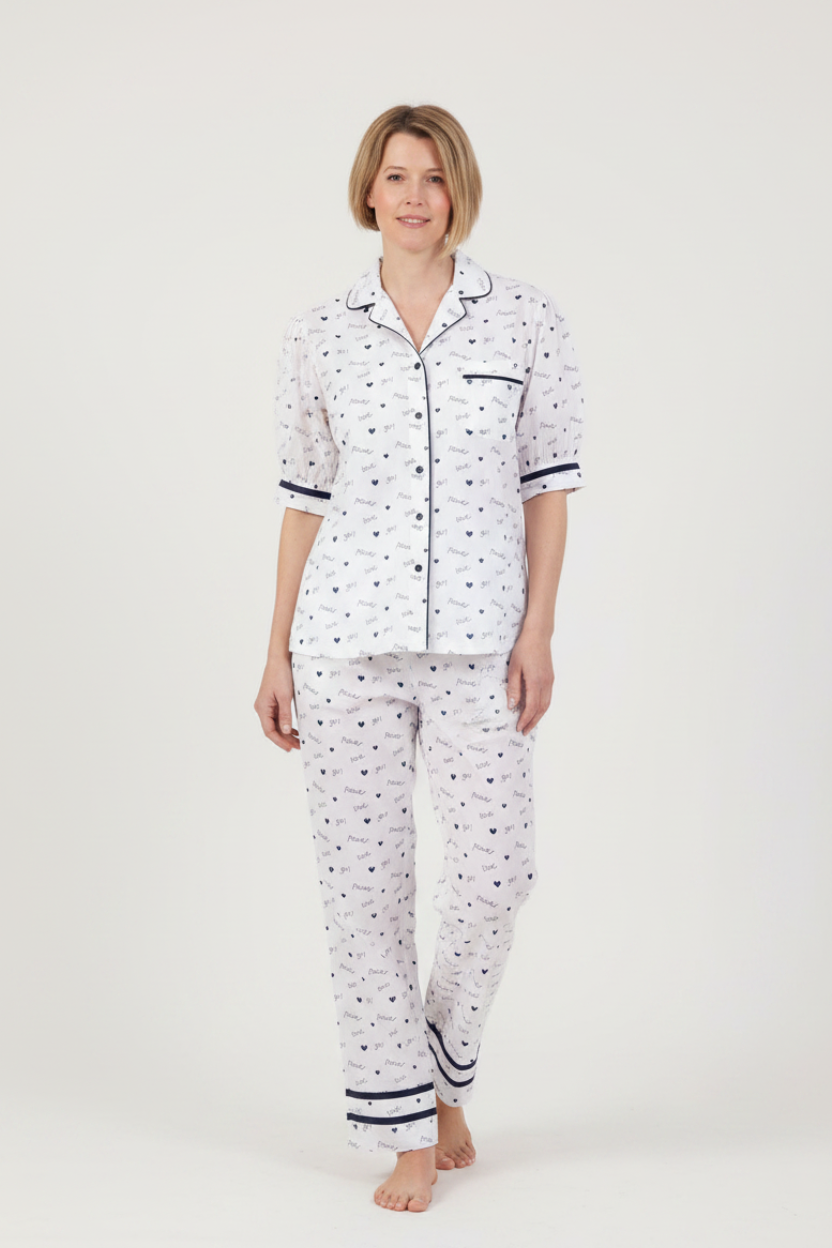 A woman modeling a two-piece pajama set featuring a white base with a small navy blue pattern, a short-sleeve button-up top, and matching long pants, set against a plain light gray studio background.