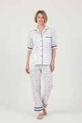 A woman modeling a two-piece pajama set featuring a white base with a small navy blue pattern, a short-sleeve button-up top, and matching long pants, set against a plain light gray studio background.