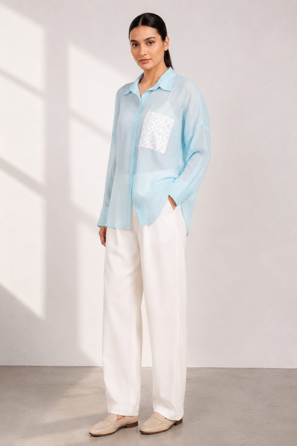 A woman modeling a light blue sheer long-sleeve button-up shirt with a white textured chest pocket, paired with white wide-leg trousers and beige loafers, standing in a brightly lit studio.