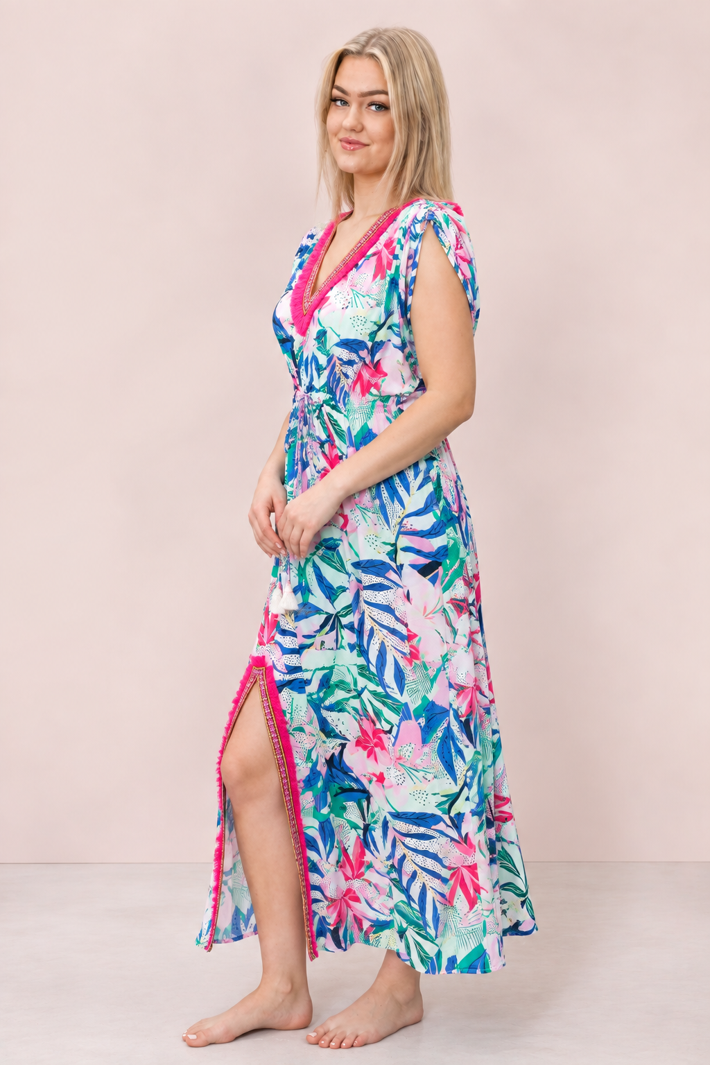 A woman modeling a vibrant tropical print midi dress with a pink trim, standing against a solid pale pink studio background.