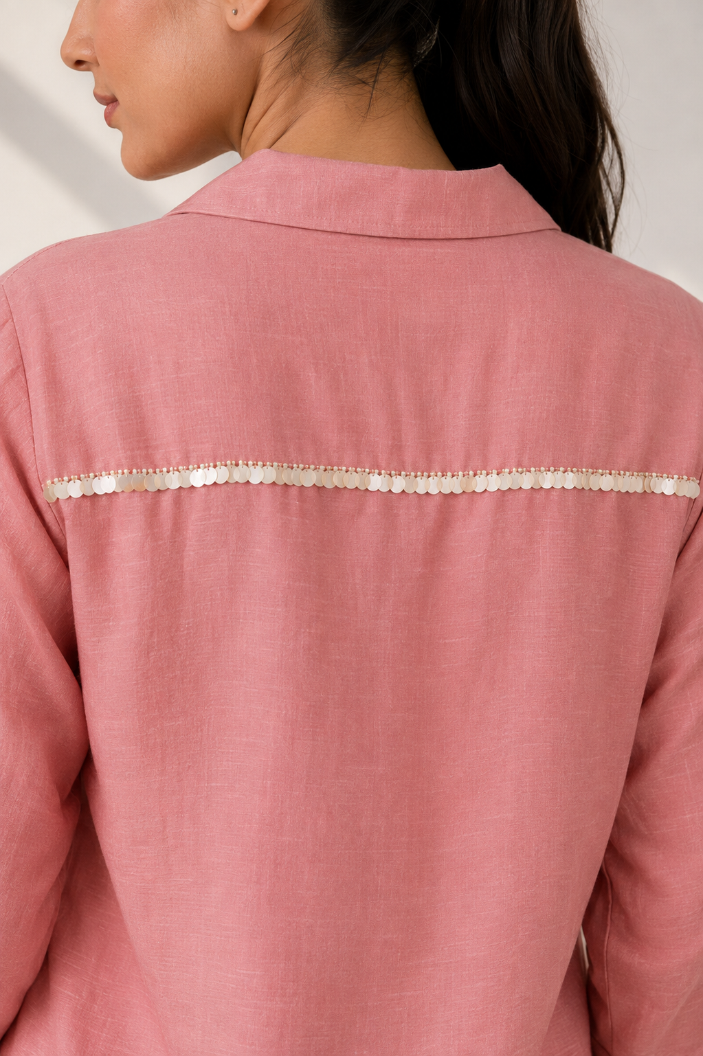 A close-up back view of a woman wearing a dusty pink linen shirt with a horizontal band of white sequins across the upper back.