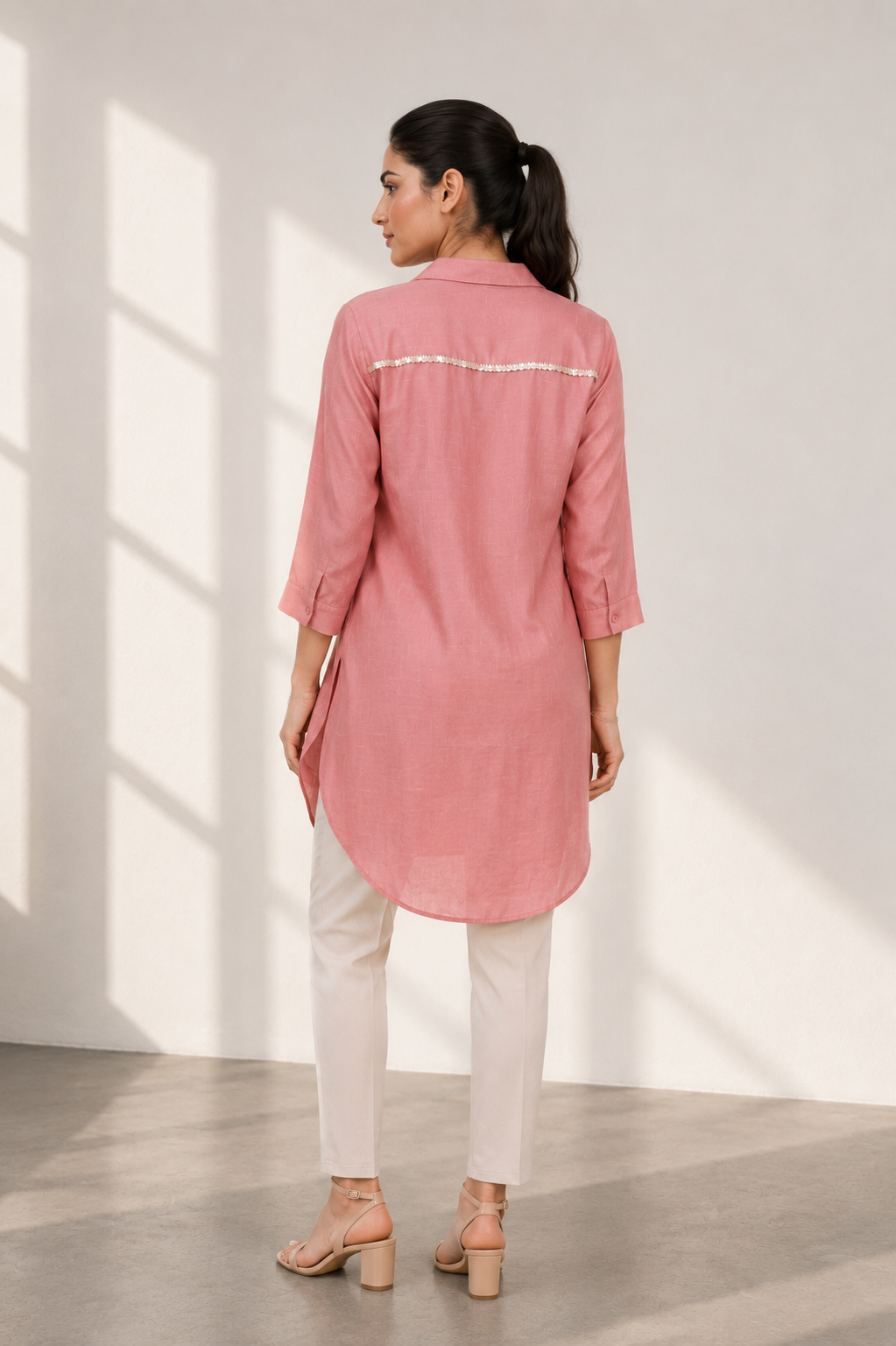 A woman is shown from the back wearing a dusty pink long kurta with white pants and beige block-heel sandals, standing in a brightly lit studio with window shadows.