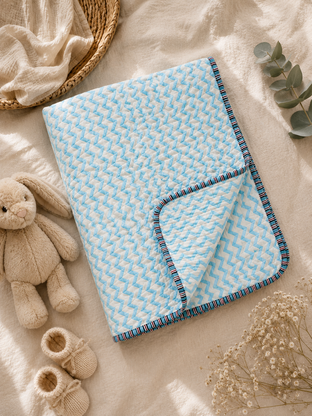 A light blue and white patterned baby blanket with a striped trim, styled in a flat lay with a plush bunny, baby booties, dried flowers, and a woven basket on a soft cream fabric background.