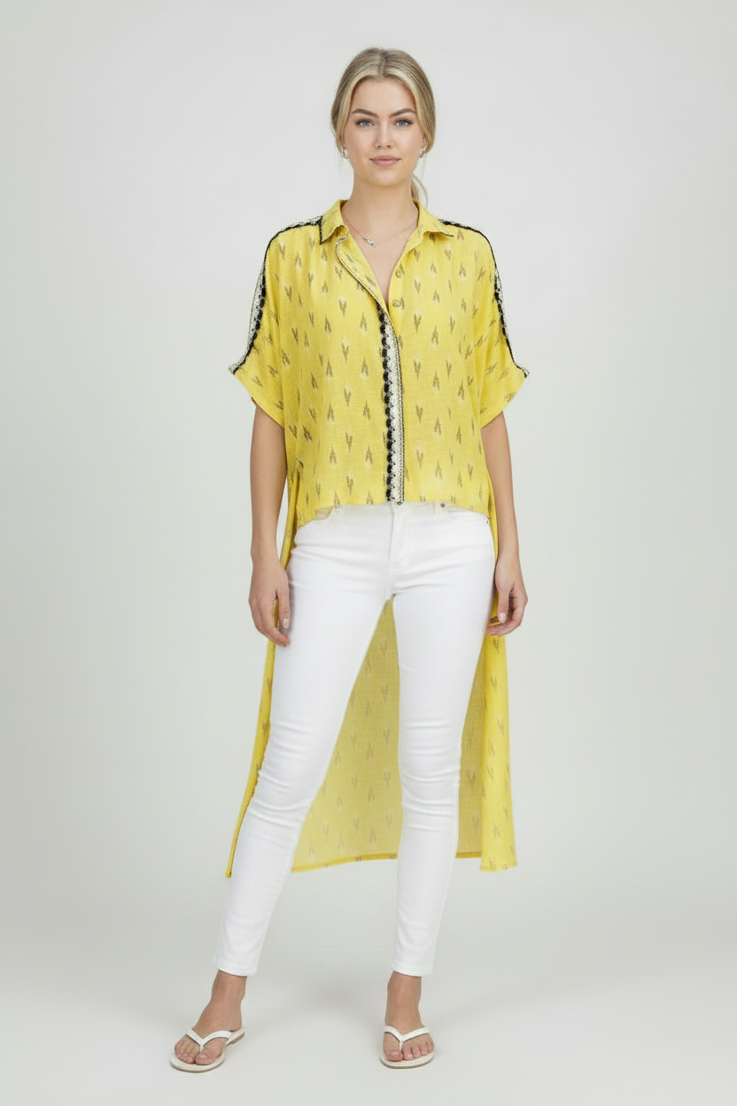 A woman modeling a yellow short-sleeved button-up shirt with a black and white geometric pattern, styled with black pants and black slip-on shoes against a light gray studio background.