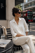 A woman wearing a white long-sleeve collared shirt and matching white trousers sits on a black outdoor cafe chair in a city setting. A red double-decker bus is visible in the background, along with a table holding a newspaper and a glass of water.