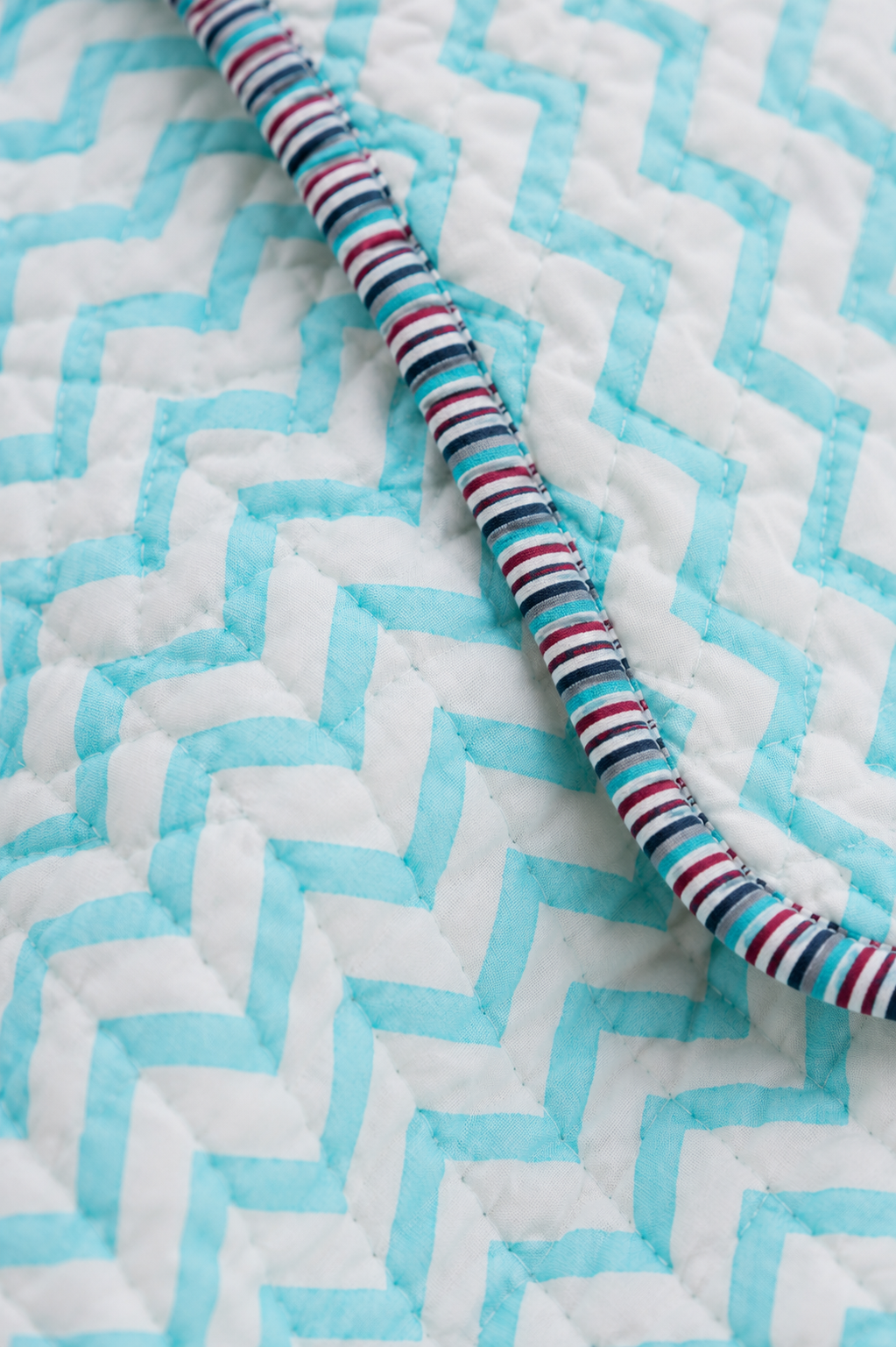 A close-up of a quilted blanket with a white and light blue chevron pattern, featuring a striped fabric trim in red, blue, white, and black running diagonally across the frame.