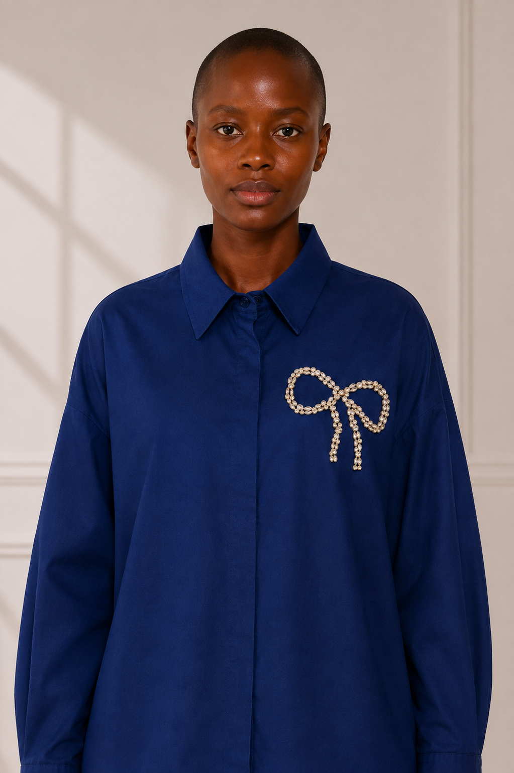 A model wearing a rnavy blue long-sleeve button-up shirt with a bow-shaped embellishment on the left chest, set against a neutral cream-colored paneled wall.