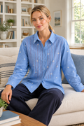 A woman sits on a light beige sofa in a bright living room, wearing a light blue long-sleeve button-up shirt with vertical embroidered stripes and small embroidered motifs, paired with dark navy pants.