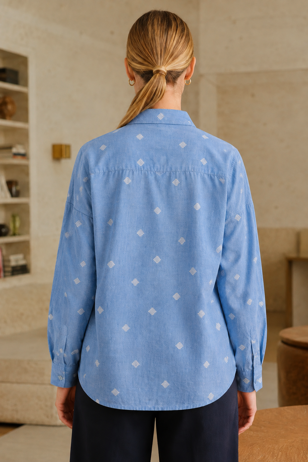 A woman viewed from behind wearing a light blue long-sleeve button-up shirt with a repeating small white diamond pattern, paired with dark navy pants in a softly lit indoor setting.