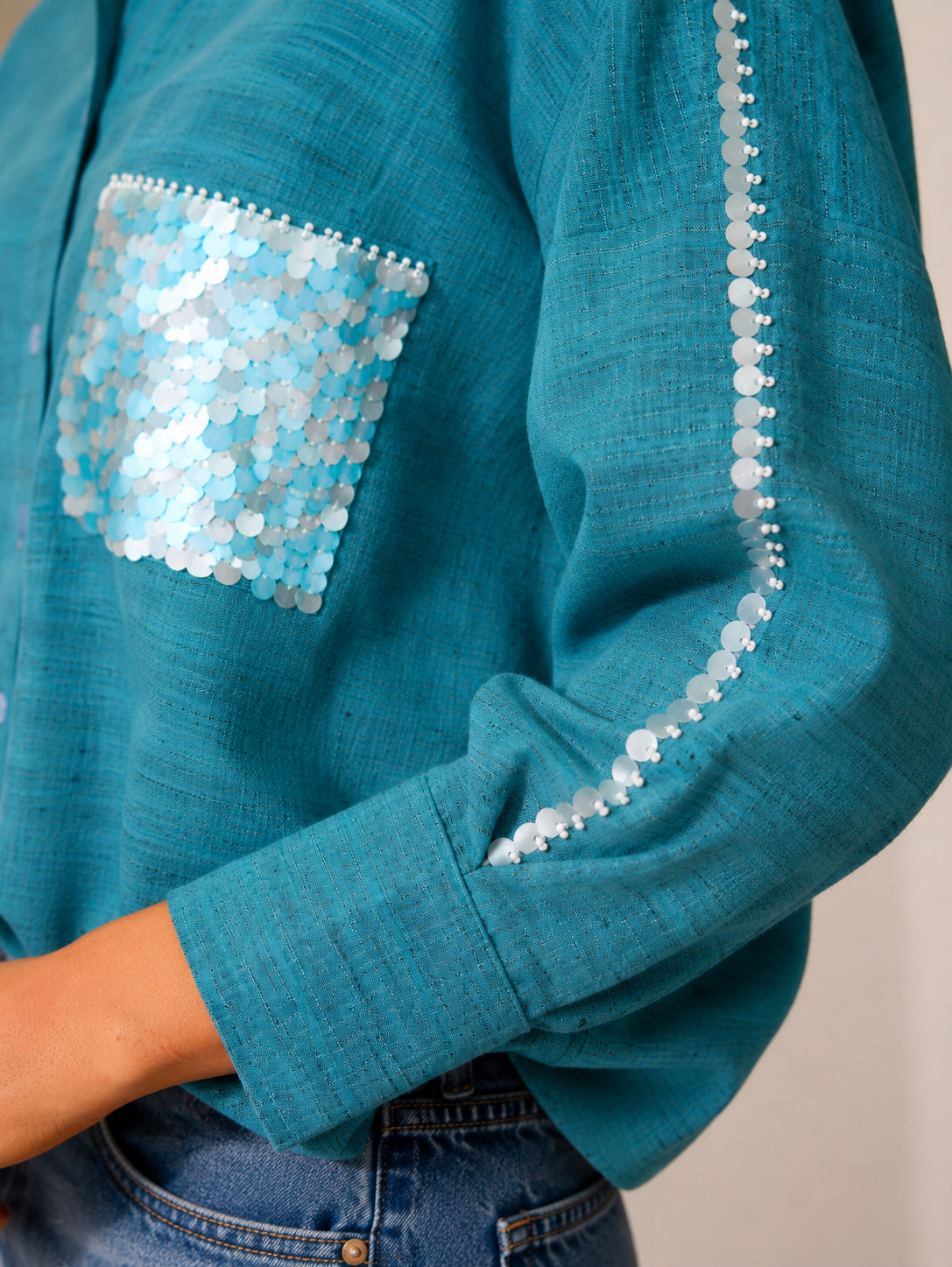 A close-up of a teal blue-green shirt with a sequined chest pocket and matching sequin trim down the sleeve, worn with blue jeans.